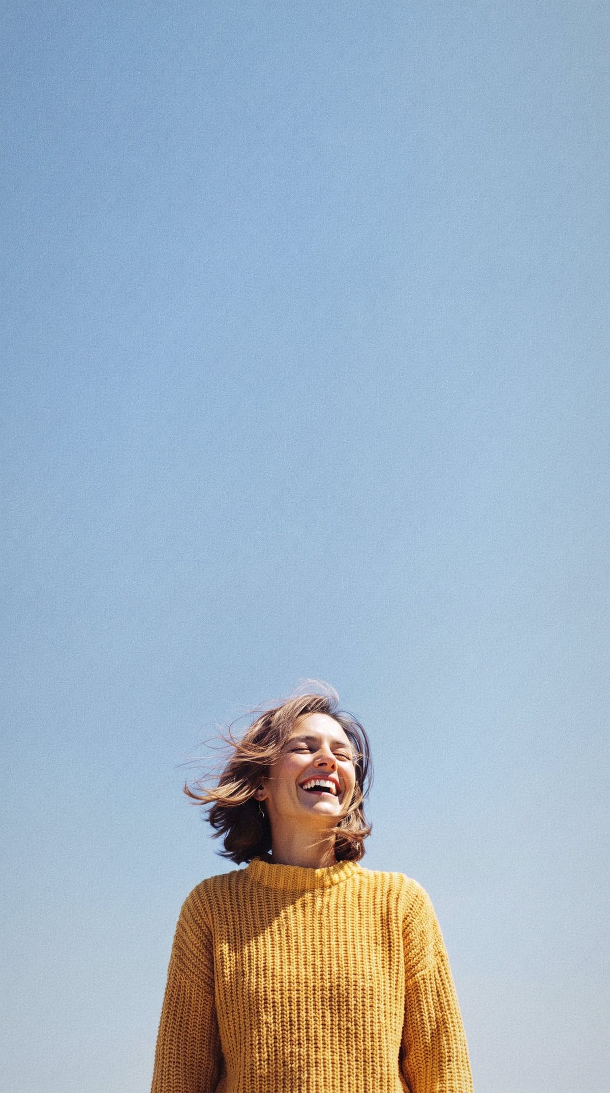 A woman with windswept brown hair laughing in the sun against a clear blue sky.