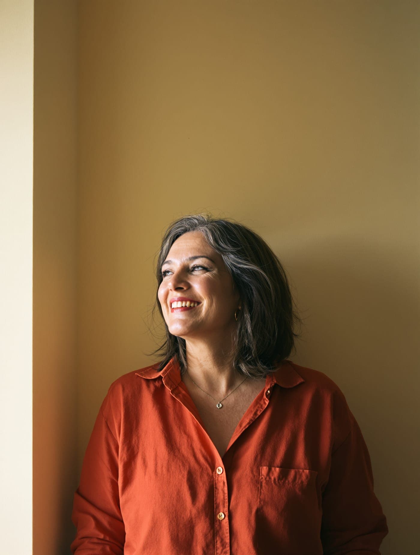 A woman in a red shirt smiling and looking away, against a warm yellow wall.