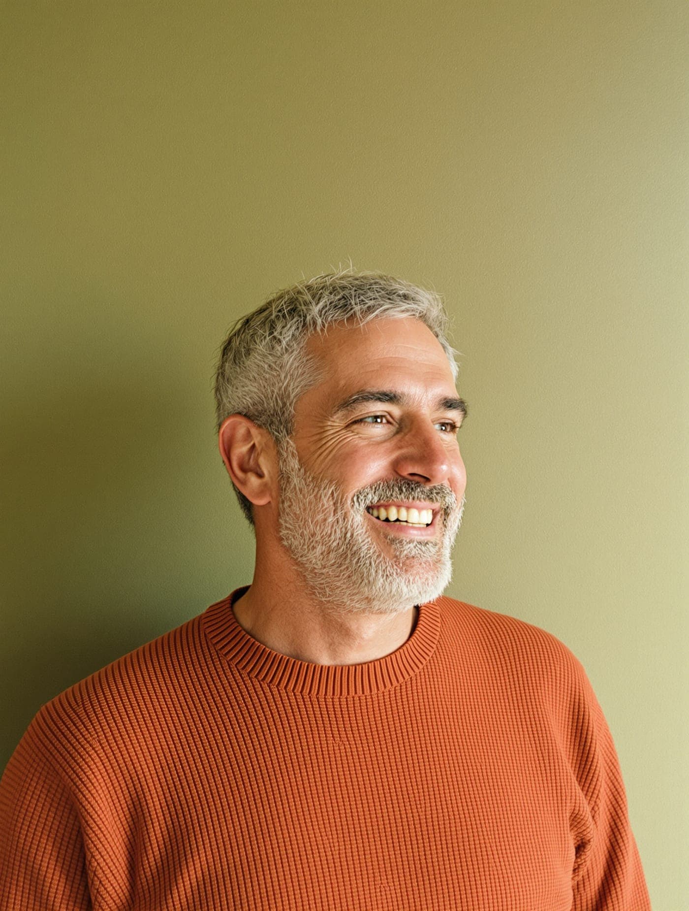A man with a grey beard in an orange sweater, smiling.